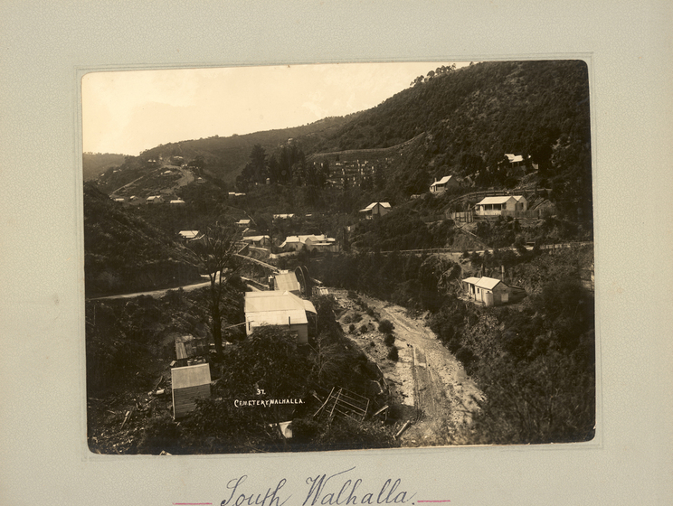 Black and white landscape photograph of a dirt road running at the base of a shallow gully, with some buildings on the hillside either side. Tree-covered hills surround. Text on the surrounding mount board reads, 'South Walhalla'. 
