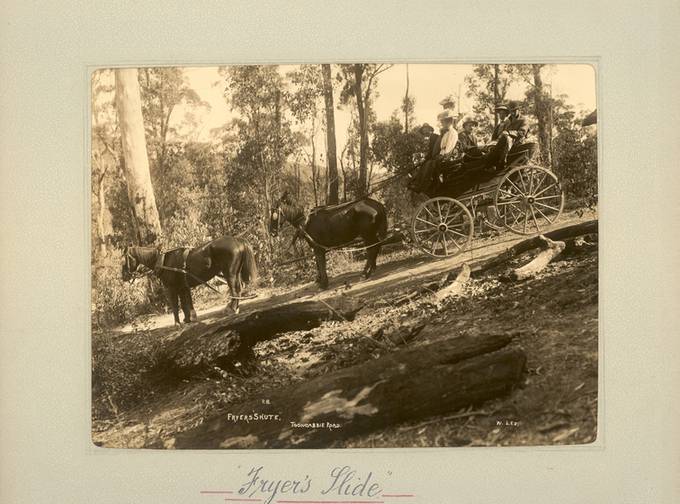 Black and white landscape photograph of two horses pulling a cart or buggy with five men and women seated in it. The surrounding landscape is bushland, and the dirt path the horses and baggy are on sloped on an angle. Text on the surrounding mount board reads, 'Fryer's Slide'.