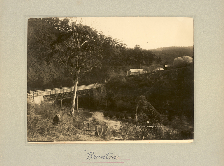 Black and white landscape photograph of a small timber bridge crossing a creek. In the distance behind trees buildings can be seen. Text on the surrounding mount board reads, "Brunton". 