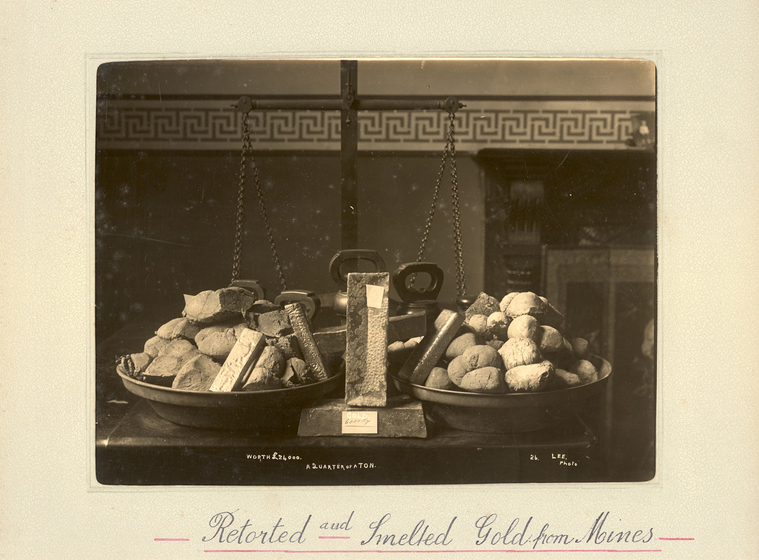 Black and white landscape photograph of a set of metal scales, with a circular pan or plate on each side holding a pile of rocks or stones. The panes are sitting on a table, and the scales are not currently balancing. Text on the surrounding mount board reads, 'retorted and smelted gold from mines'