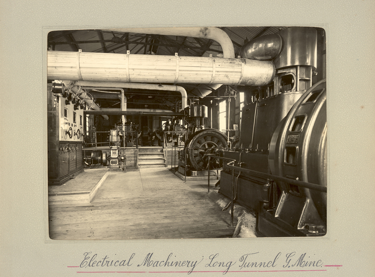 Black and white landscape photograph showing inside a factor with timber floors, and large machines across the factory floor. A large white pipe runs across the top of the room. Text on the surrounding mount board reads, 'Electrical Machinery Long Tunnel G. Mine'