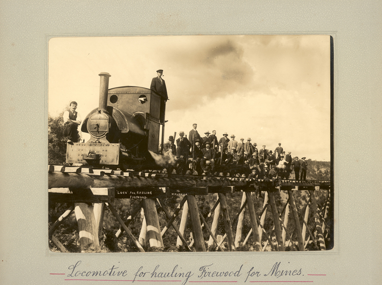 Black and white photograph of an elevated rail bridge, with a train engine and a large group of men standing on the track behind. Text on the surrounding mount board reads 'Locomotive for hauling Firewood for Mines'