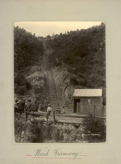 Black and white portrait photograph of train tracks running up a hill side. In the foreground is a group of people seated on a timber fence looking back at the train line. There is a small wooden hat at the base of the hill. Text on the surrounding mount board reads 'Wood Tramway'