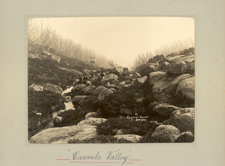 Black and white landscape photograph of rocks in a shallow dip in the terrain, with a small stream running through. There is a group of people seated in the distance on some of the rocks. Text on the surrounding mount board reads 'Cascade Valley'. 