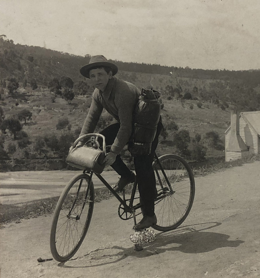 Man with hat riding a bicycle along a road through countryside past a house, trees and hills in background. Man wearing a hat and has a bag slung over his left hip and another across the handlebars of the bicycle.