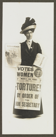 Photograph of woman holding newspaper titled "Votes for women" in right hand, another folded over left arm, left hand holding a "Votes for Women" newspaper billboard sheet with headline "Torture! By order of the Home Secretary". Woman is wearing a hat and a long dress.