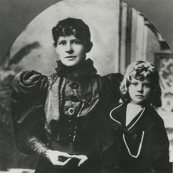 Black and white studio portrait of a woman in an elaborate Victorian dress holding the hand of a blonde boy.