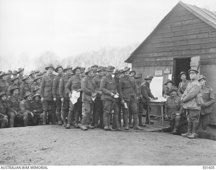 Photograph showing soldiers in uniform, some holding pieces of paper lining up in front of a table outside a hut. Soldier at the front of the line appears to be writing. An officer and some other soldiers are standing in front of the hut. All are looking at the camera.