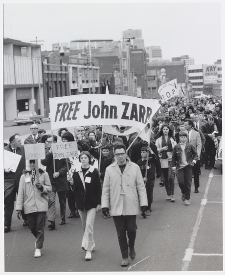 Photograph of a crowd of people marching along a city street, several carrying placards and a large banner saying "Free John Zarb".