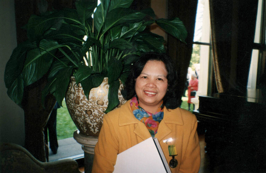MIddle aged woman wearing an orange blazer, with a medal on her chest pocket, and a coloured scarf. She holds a folder and stands in front of a large potted plant