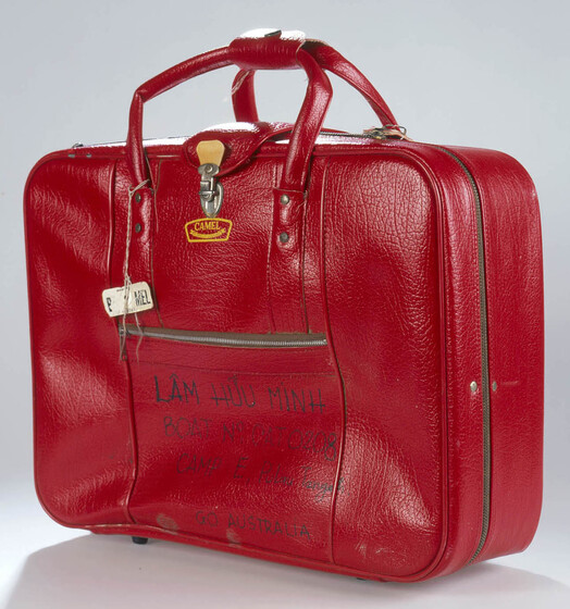 Red vinyl small suitcase with zip on size and two carry handles. Text on side pocket in texta reads 'LAM HUU MINH' with a boat number and camp name, with words 'go Australia' beneath.