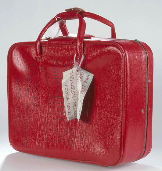 Red vinyl small suitcase with two carry handles. Text on side in texta reads 'LAM HUU MINH' with a boat number and camp name, with words 'go Australia' beneath. A white tag hangs from handles.