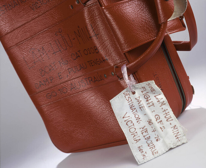 Red vinyl small suitcase standing on its side with two carry handles. Text on side in texta reads 'LAM HUU MINH' with a boat number and camp name, with words 'go Australia' beneath. A white tag hangs from handles.
