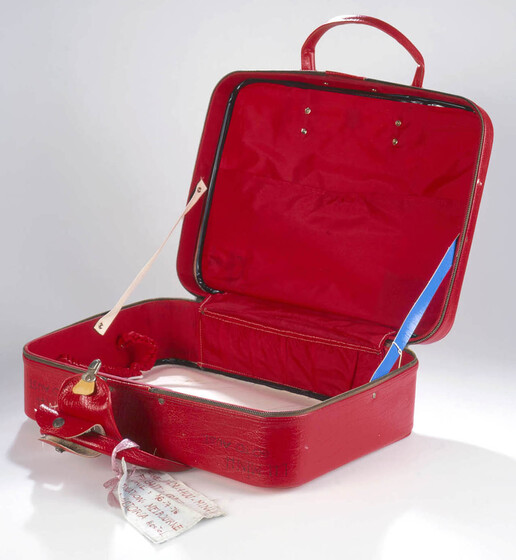 Empty red vinyl small suitcase with lid open. A white tag hangs from from of the carry handles.
