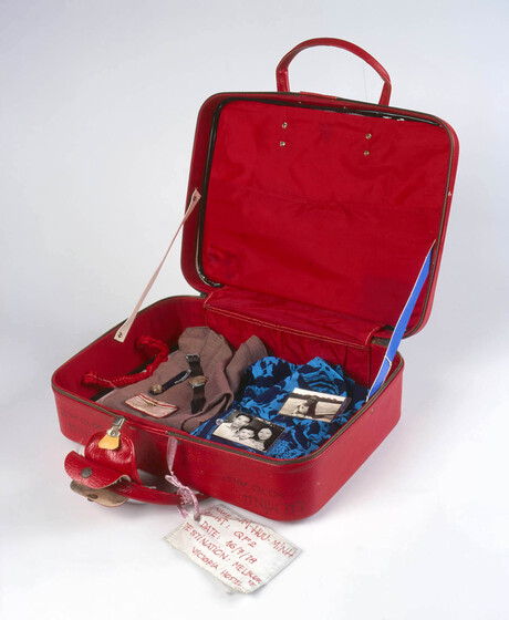 Red vinyl small suitcase with lid open containing clothing and other items. A white tag hangs from from of the carry handles.