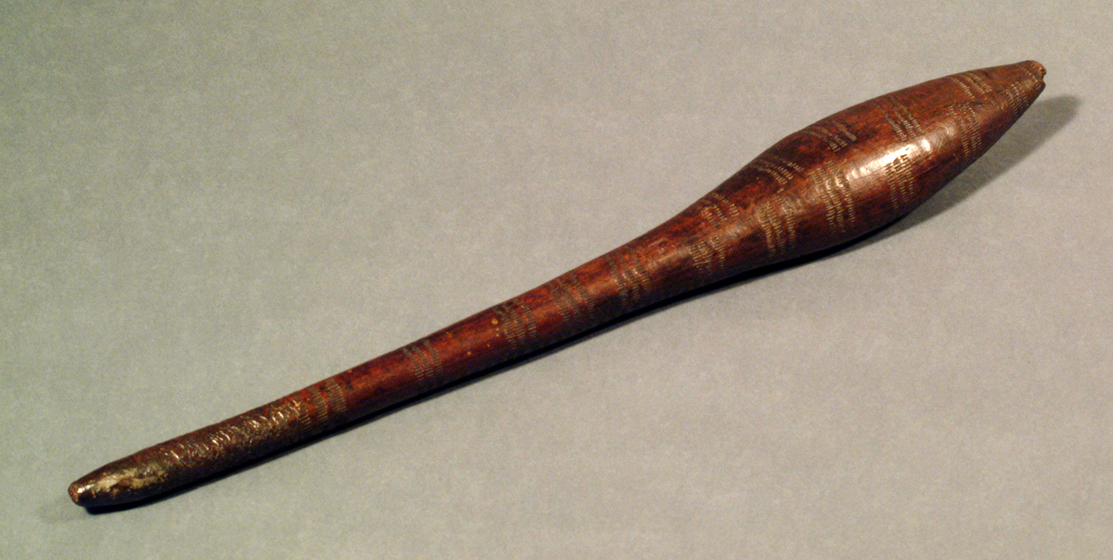 Thin, wooden club with carved designs covering the surface. One end of the club is wider than the handle-end.