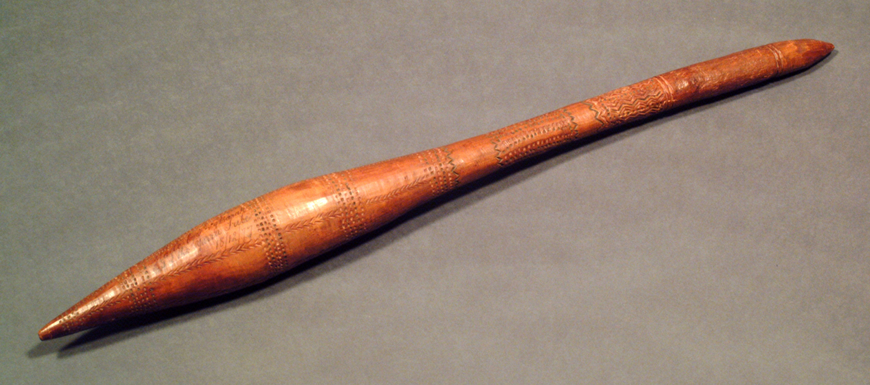 Thin, wooden club with carved designs covering the surface. One end of the club is wider than the handle-end.