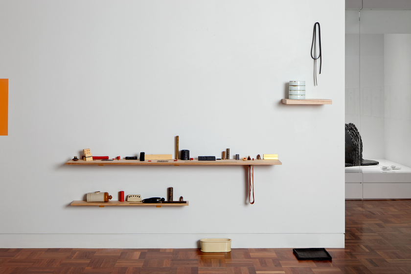 A white wall on which are three wooden floating shelves. The largest is in the middle and has a loop of fabric hanging from its right hand side. The smallest is to the top right with a loop of material hanging above it, at the bottom is the middle-sized shelf. On these shelves are an assortment of ceramic pieces which look like household items arranged in a particular way. with a mat and small metal container on the floor beneath. The wall and display is in a gallery with parquetry floor, and other works can be seen in the distance and sides. 