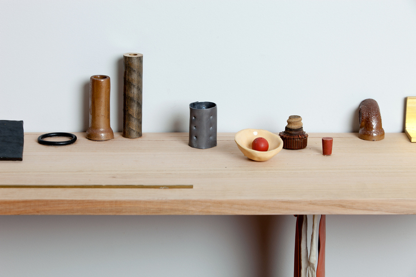 Close up view of a wooden floating shelf on a white wall, featuring a range of ceramics that look like household items arranged in a specific way, including a small bowl with a red ball in it, cylindrical handles and pipe ends, and a rubber ring.