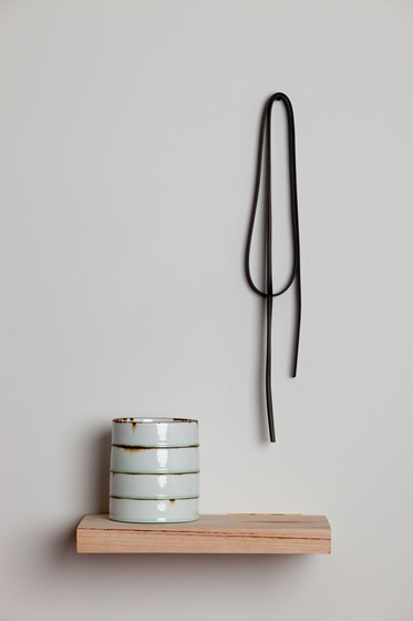 Close up view of a small wooden floating shelf on a white wall. On this shelf is a white ceramic circular container, and above this hangs a loop of material. 