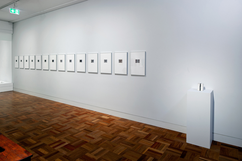 A white wall featuring a row of fourteen small artworks in white frames all hung in a row. On the right is a white rectangle block with a ceramic item on top. The floor of the gallery is parquetry.