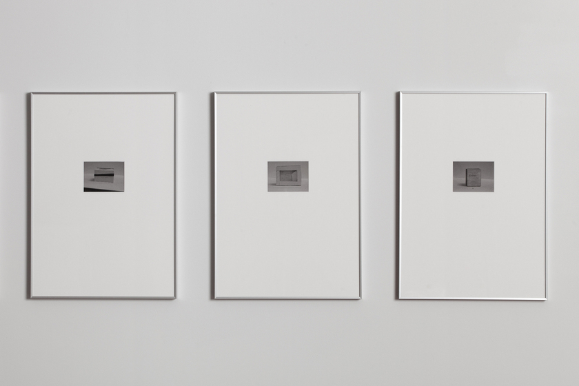 Three white frames containing small photographs of boxes, hung in a row on a white wall.