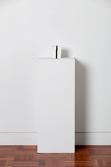 White box sitting on a white rectangle box, with a black band wrapped over the top and front side. The box is sitting on a parquetry floor in front of a white wall.