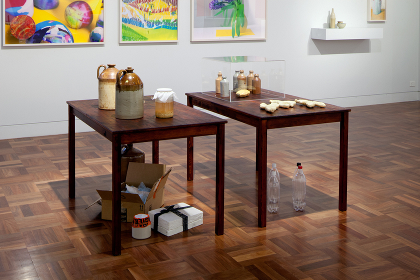 Two tables placed in the middle of a room with white walls, standing on a parquetry floor. On the tables are ceramic jugs, and other small items. Underneath the tables are plastic bottles, and cardboard boxes. On the wall behind are hung other artworks.