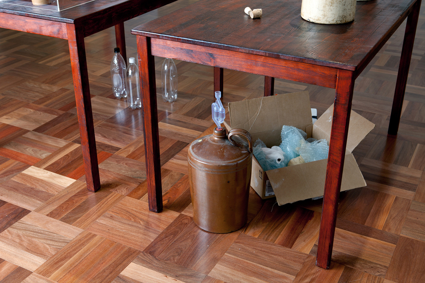 Close up view of two wooden tables standing on a parquetry floor. Underneath the tables are plastic bottles, and cardboard boxes.