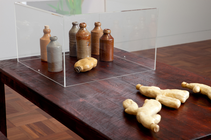A wooden table standing on a parquetry floor. On the table are ceramic jugs under a plastic transparent through box, and other small items. On the wall behind are hung other artworks.