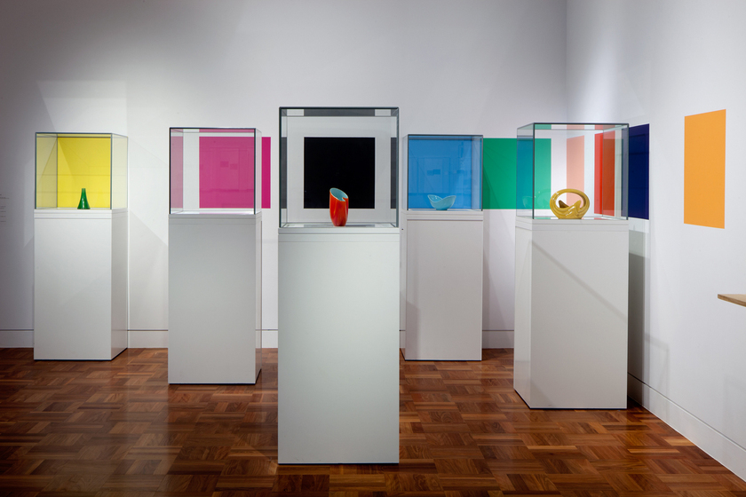 Four ceramic artworks displayed individually on four white columns and covered with glass boxes. There is a fifth white column with no artwork inside. On the white wall behind is multiple different coloured squares.