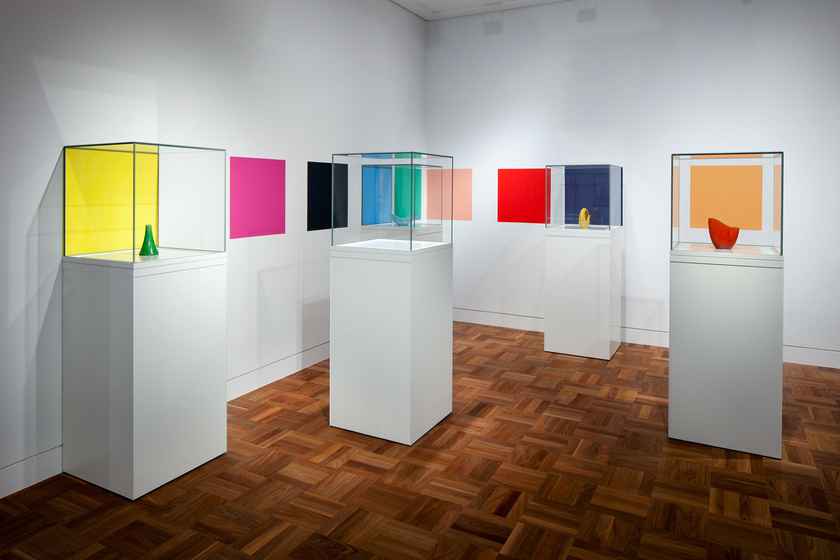 Four ceramic artworks displayed individually on four white columns and covered with glass boxes. There is a fifth white column with no artwork inside. On the white wall behind is multiple different coloured squares.