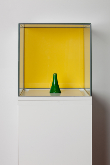 A green ceramic flask displayed on a white column under a glass box, with a yellow square on the wall behind.