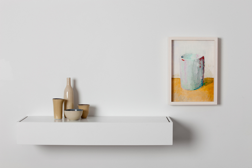 A white shelf with four tan coloured ceramic pieces displayed on it, a bowl, two cups and a bottle. To the right is a painting of a blue cup on a yellow surface in a white frame.