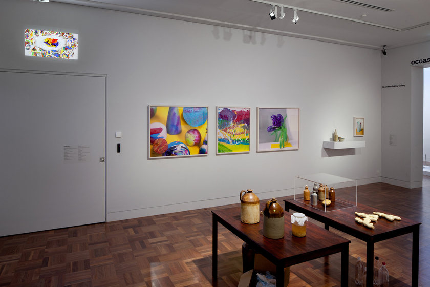 A room with white walls and parquetry floor, with three coloured framed artworks hanging on the wall, two wooden tables with various ceramic bottles on the surface, and a small shelf with ceramic items next to a small painting.