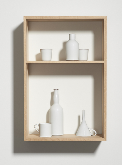 A timber shelf made with two square boxes hung on a white wall to create shelves. Inside the bottom box is a white cup, bottle and funnel with a handle. Inside the top box are two white cups and a bottle.