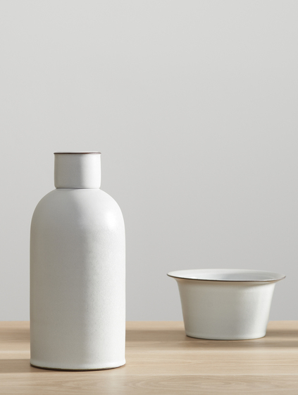 A white bottle on the left and white bowl on the right, sitting on a timber shelf.