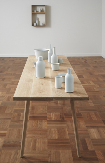 A timber table standing on a parquetry floor. On the table is a series of white bottles, cups and bowls. Behind is a white wall with two wooden boxes forming two shelves holding white bottles and cups.