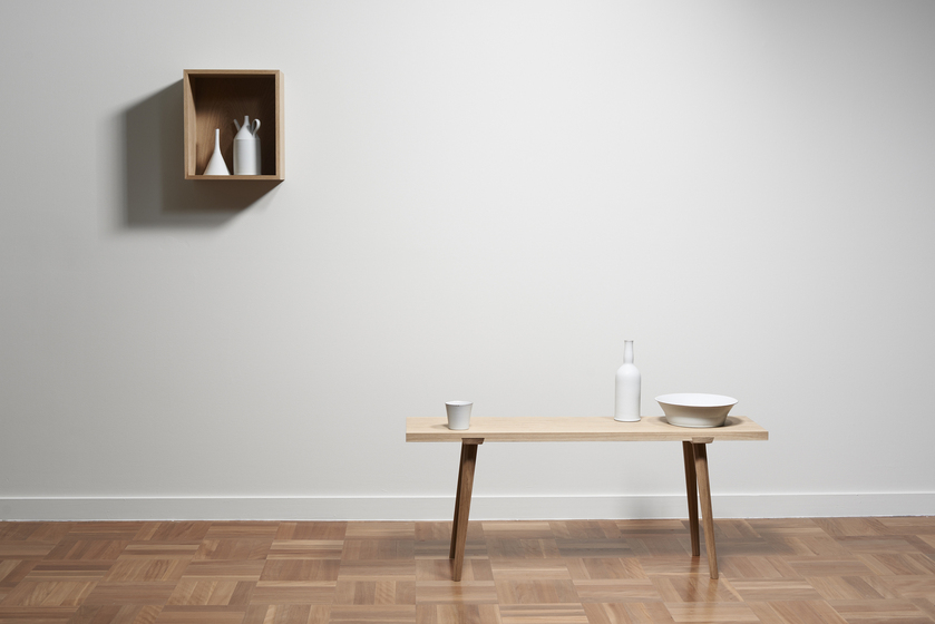A timber box hung on a white wall to create a shelf. Inside the box is a white flask and white jug with a spout and handle. To the right is a wooden table sitting on a parquetry floor. On the table is a white cup, bottle and bowl.