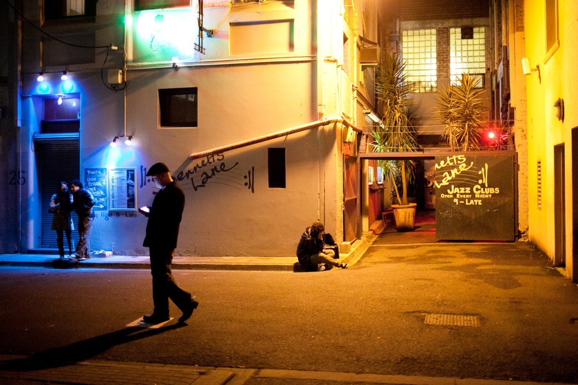 Colour photograph of a man wearing a black jacket and cap, walking through a laneway at night. The building behind is lit with blue and green lights, and two people stand under the blue light in front of a door. Written in black grafitti on the building is the words 'Bennets Lane'. Written again on a black board is the words 'Lane' with 'Jazz Clubs' written underneath. 