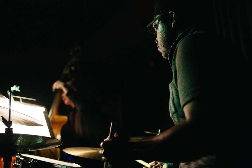 Colour close up photograph of a man in a green shirt wearing glasses seated at a drum set. Only the cymbals are seen. In the distance is a person with a double bass.