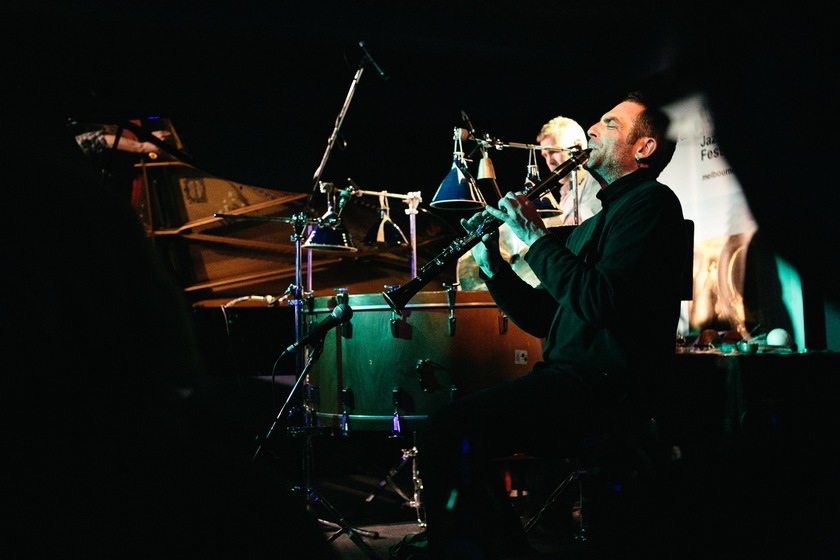 Colour photograph of a man seated playing a clarinet with a microphone in front. Behind is a person standing at a large drum. 