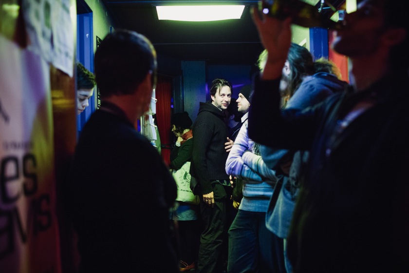 Colour photograph of a corridor with papers on the walls and blue doorframes, crowded with people. An out of focus person in the foreground drinks from a bottle. At the end of the corridor is a man in a black hoodie looking back at the viewer. I