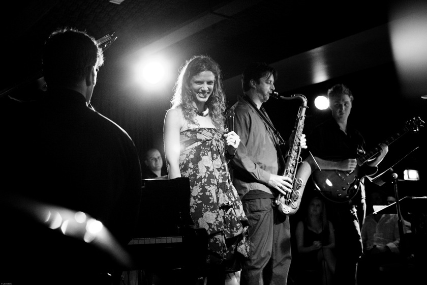 Black and white photograph of a woman in a floral strapless dress holding a microphone. To the right is a man playing a saxaphone in and a man playing a bass guitar. In the foreground to the left is the back of a person playing a piano.