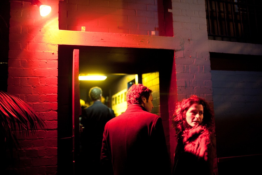 Colour photograph of a man and a woman walking through an open door of a brick building into a yellow-lit corridor. A red light shines on them, and the building their walking into.