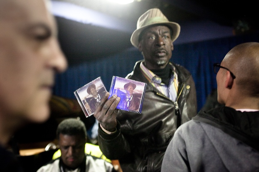 Colour photograph of a man in a hat holding up two CD cases with one hand. A man stands in front of him looking at this face, and the face of another man is out of focus in the foreground. Blue curtains are seen behind.