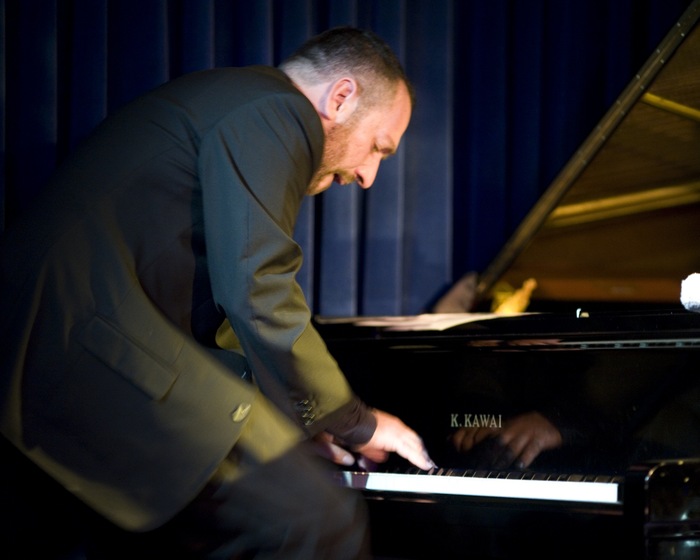 Colour photograph of a man in a suit jacket standing hunched over a piano with the brand name 'K. Kawai' seen, playing the keys with blue curtains behind..