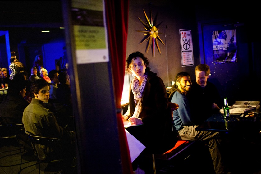 Colour photograph of a woman with a scarf around her neck, seated against a call at a small table with papers on it. Behind her on the wall is a large metal sun-like wall hanging, and two men seated on a bench with a bottle on the table in front of them. In the foreground out of focus is a pillar. To the left of the pillar can be seen a crowd of seated people.