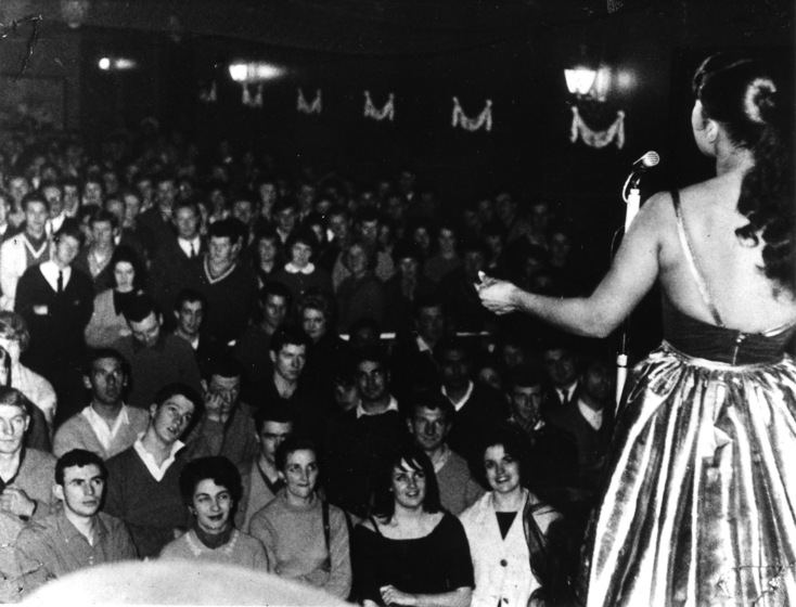 Black and white photograph of a woman wearing a shiny skirt, standing in front of a microphone facing away from the camera towards a large audience of people