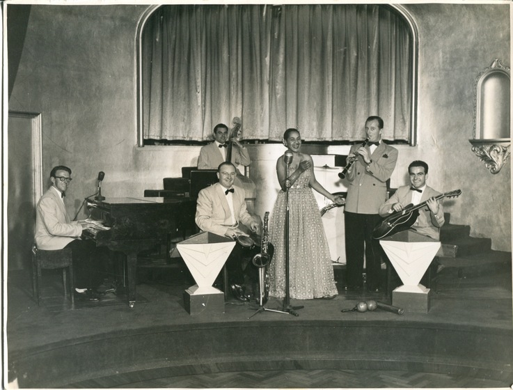 Black and white photograph of a woman standing centre of a stage wearing a long dress behind a microphone. Surrounding her are five men in light coloured suit jacks, playing different instruments, including a grand piano, double base, saxophone, clarinet, and guitar. 